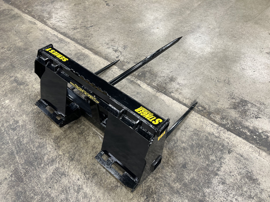 Heavy-duty bale spear by Stinger Attachments mounted on skid steer – designed for easy transport and stacking of large hay bales.
